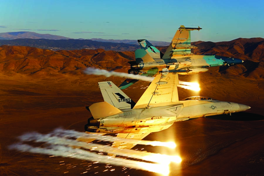 A U.S. Navy Naval Aviation Warfare Development Command Boeing F/A-18F Super Hornet and a camouflaged F/A-18C Legacy Hornet maneuver while dispensing flares over the Nevada desert. Although the Super Hornet is 20-25% larger than the legacy, the easiest way to externally tell them apart is by the square intakes on the Super versus the radius-shaped intakes on the Legacy. Other clues include the Rhino’s larger leading edge extension and the leading edge of the wing. Unlike the Legacy, the Rhino has a step where the wing folds.